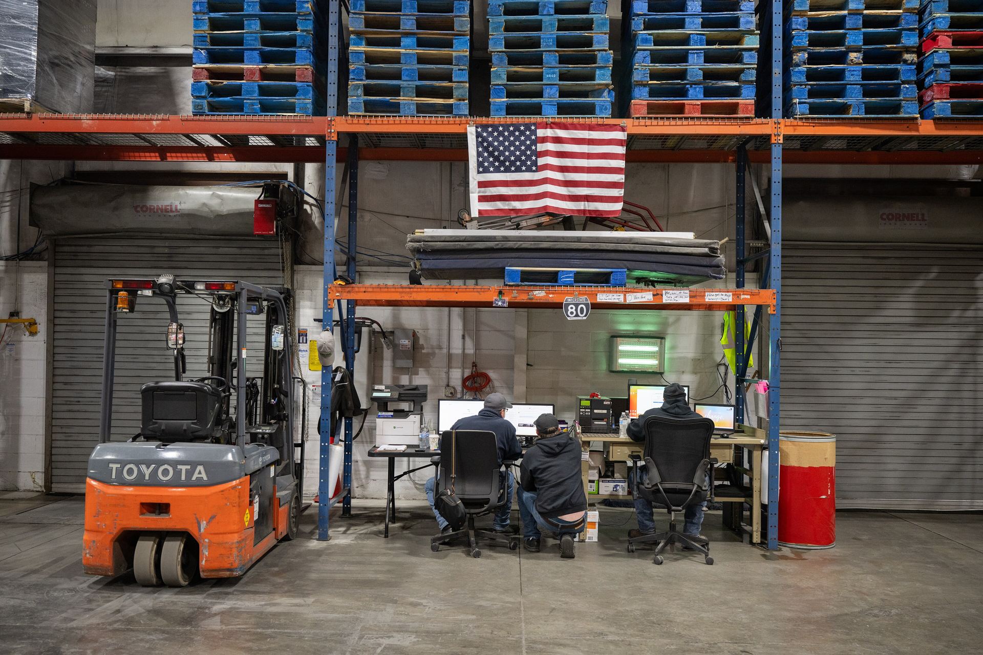 Three people at computers in a warehouse office, with empty pallets stacked above them and a forklift beside the desk